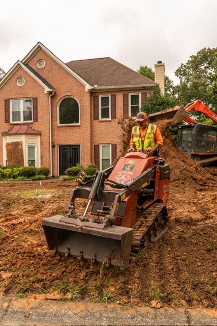 Worker operates a red mini loader in front of a brick house under construction.