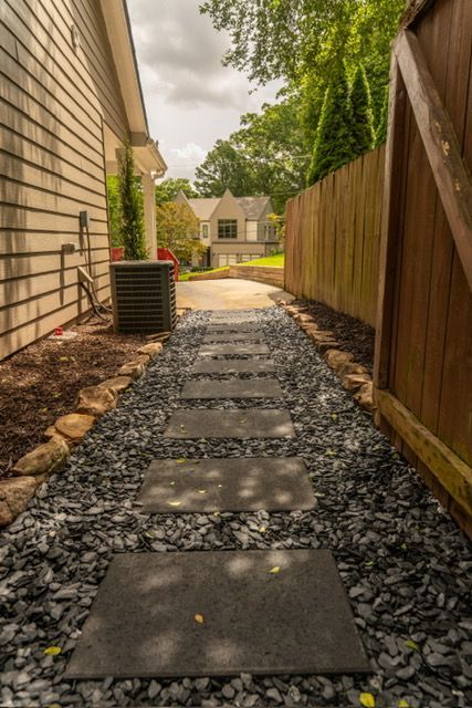 Stone stepping path beside a house leading to a suburban home, bordered by gravel, fence, and greenery.