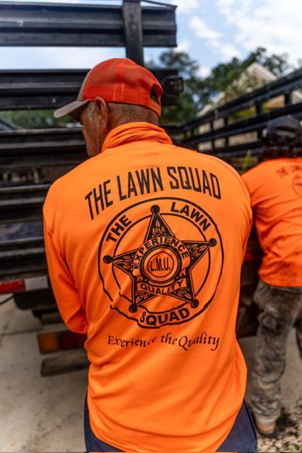 Worker in bright orange “The Lawn Squad” shirt standing by stacked black materials outdoors
