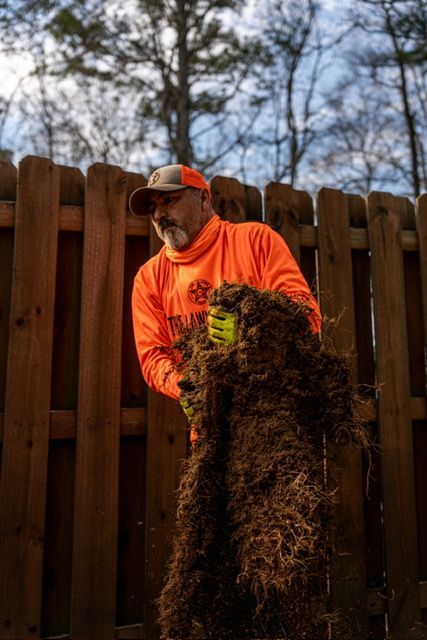 Person in a neon orange shirt and cap cleaning debris beside a wooden fence outdoors.