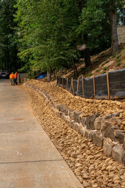 Steep roadside with rock retaining wall, trees, and workers in orange vests on a paved lane