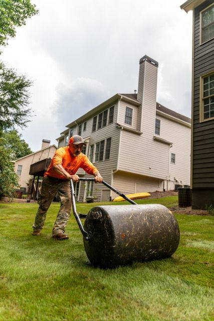 Worker in orange safety gear rolling a lawn roller on green grass beside a house.