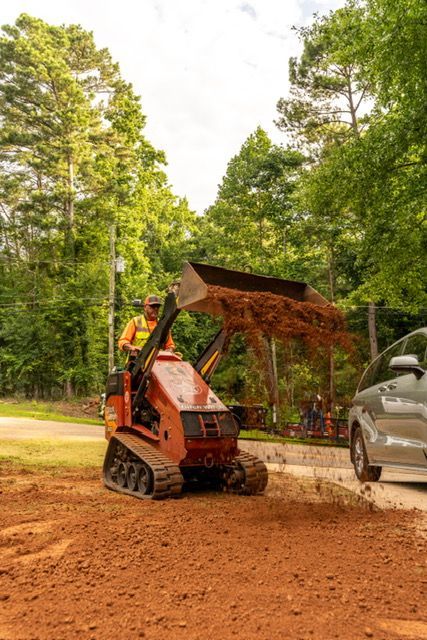 Orange skid steer loading dirt beside a gray truck on a wooded gravel drive