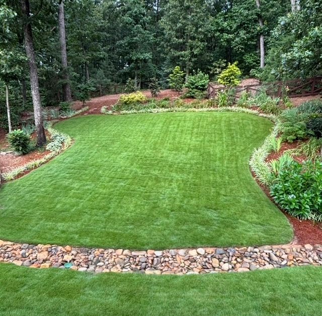 Lush green lawn bordered by red mulch, rocks, and dense trees in a landscaped garden