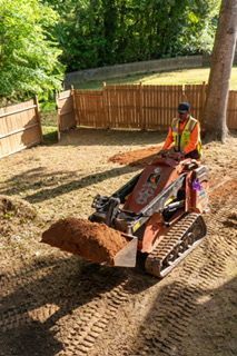 Person operating a red skid steer loader carrying dirt in a fenced outdoor yard.