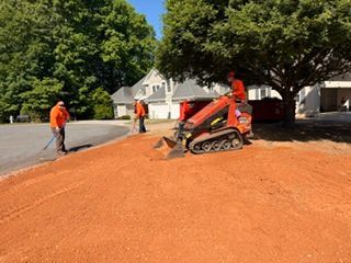 Workers in orange gear spread red mulch with a skid-steer near a tree on a sunny street.