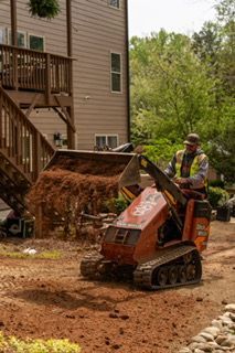 Worker operating a compact loader beside a house, moving dirt in a landscaped yard