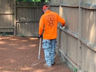 Person in orange shirt and camo pants raking leaves along a wooden fence in a backyard.