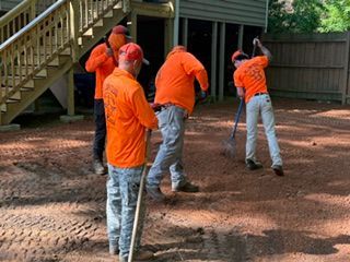 Workers in orange shirts shoveling dirt near a house and wooden stairs