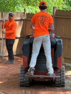 Two workers in orange shirts operating a compact skid steer in a fenced dirt yard.
