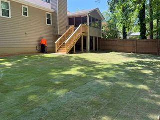 Backyard lawn with a wooden deck and stairs beside a beige house; person in orange near the wall