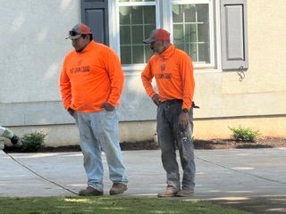 Two workers in orange shirts and caps stand on a sidewalk near a building, one holding a long tool.