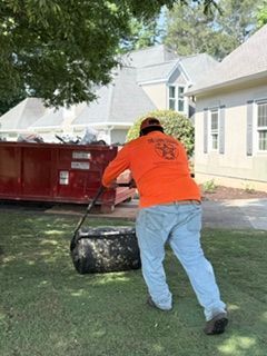 Worker in orange shirt dragging a black bin across a lawn beside a red dumpster and house
