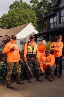 Five people in orange shirts pose on a street, one kneeling, with trees and houses in the background.