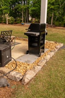 Outdoor black gas grill on a concrete patio, bordered by rocks and grass near a porch post.