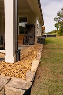 Side view of a house with a rock-lined drainage bed and grassy yard along the foundation.