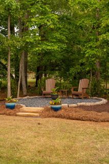 Two wooden chairs by a stone fire pit in a wooded backyard garden