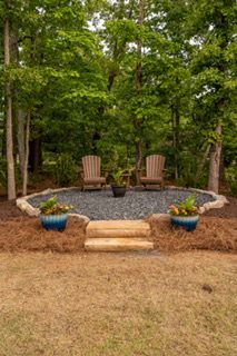 Backyard patio with two wooden chairs, a fire pit, and blue flower pots amid trees and mulch