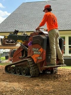 Orange-clad worker operating a tracked construction machine on a dirt site, lifting a load of soil.