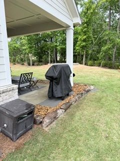 Covered grill on a small patio beside a white house, with chairs and a lawn beyond.