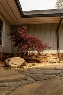 Courtyard with stone patio, tan siding, boulders, and a red-leaf shrub under an overhang