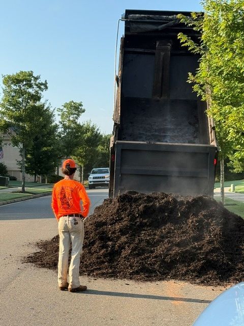 Worker in orange shirt stands by a dump truck unloading dark mulch on a street.