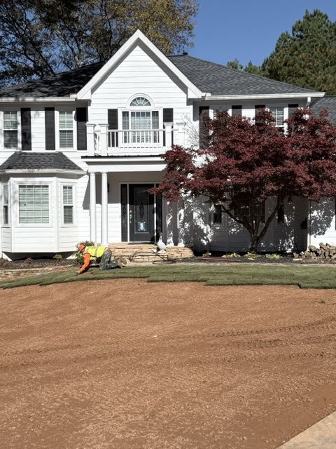 Two-story white house with new dirt front yard, workers installing landscaping, and a red-leaf tree on the right
