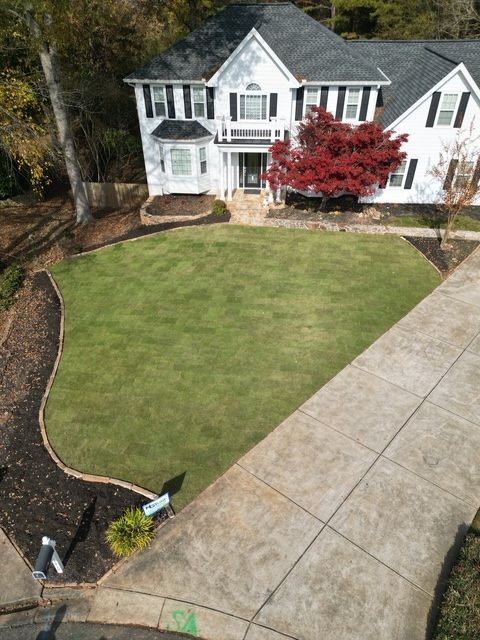 Aerial view of a suburban house with a large front lawn, driveway, and a red tree in the yard.