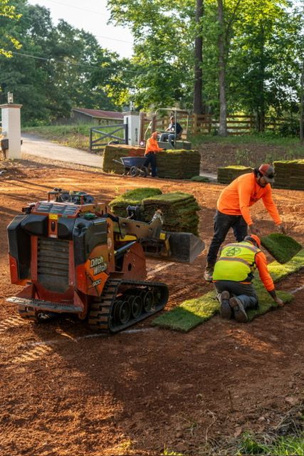 Workers in orange safety gear paving a path with a small tracked machine and tools.