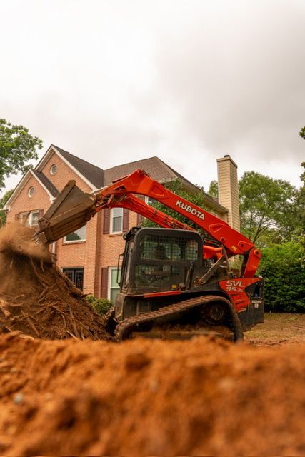 Orange excavator clearing dirt beside a brick house with a chimney and trees.