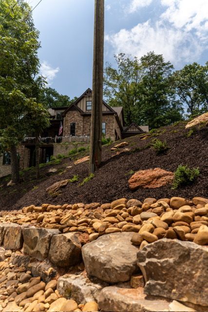 Stone house on a hillside behind a rock retaining wall, with trees and a utility pole under a cloudy sky
