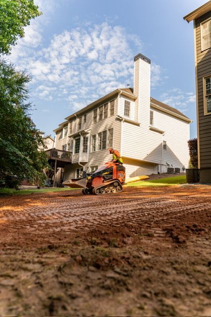 Worker operating a mini excavator on a muddy construction site beside a white house.