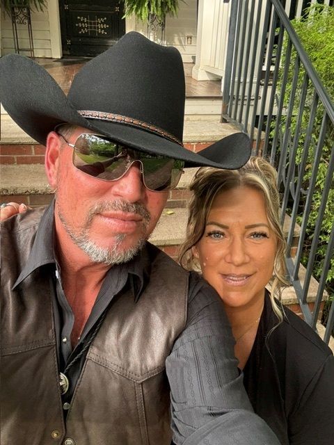 Two people posing outdoors on steps, one in a black cowboy hat and sunglasses, smiling close together.