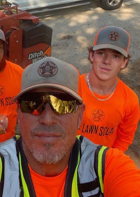 Three utility workers in orange shirts and gray caps posing outdoors in sunlight