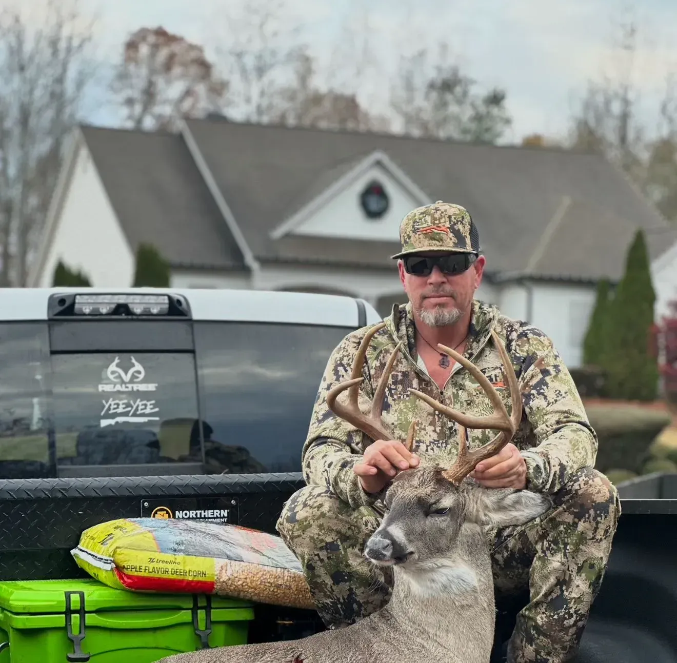Man in camouflage with a deer he hunted, posing in truck bed in front of a house.