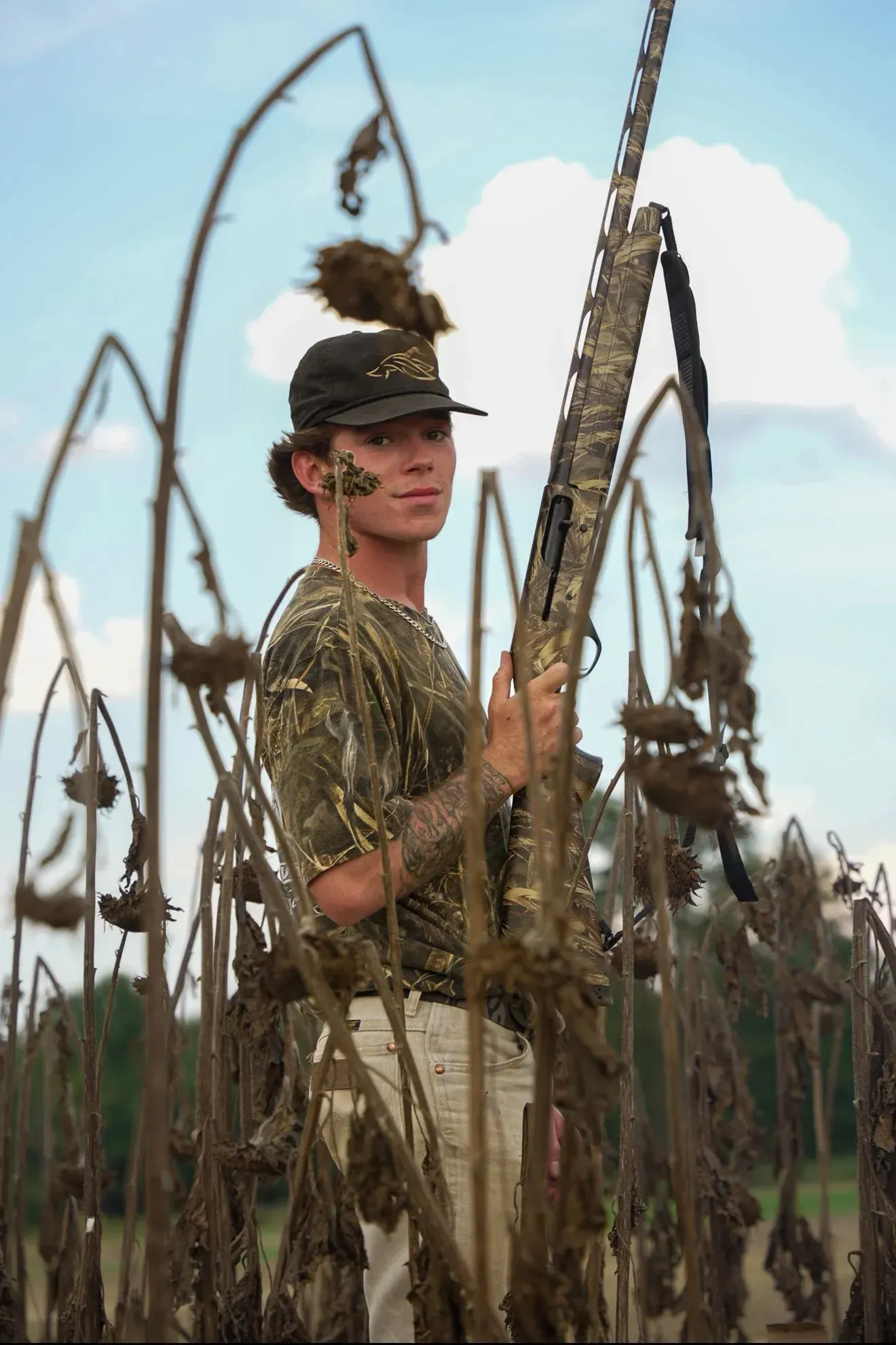 Person in camouflage gear holding a shotgun, standing in a field of dry sunflowers.