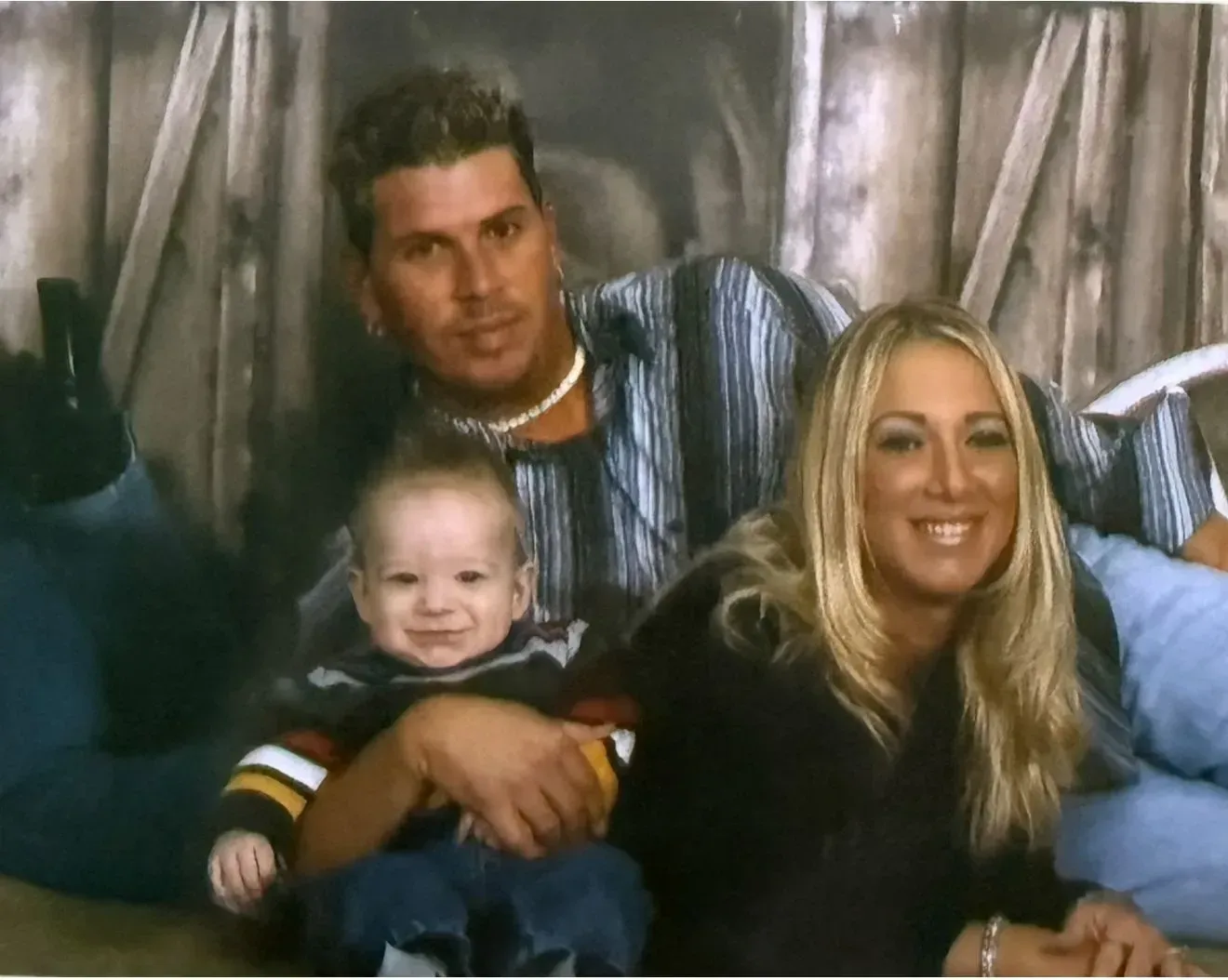 Family of three poses, smiling. Man in striped shirt, woman in black, baby in arms. Background: rustic wood.