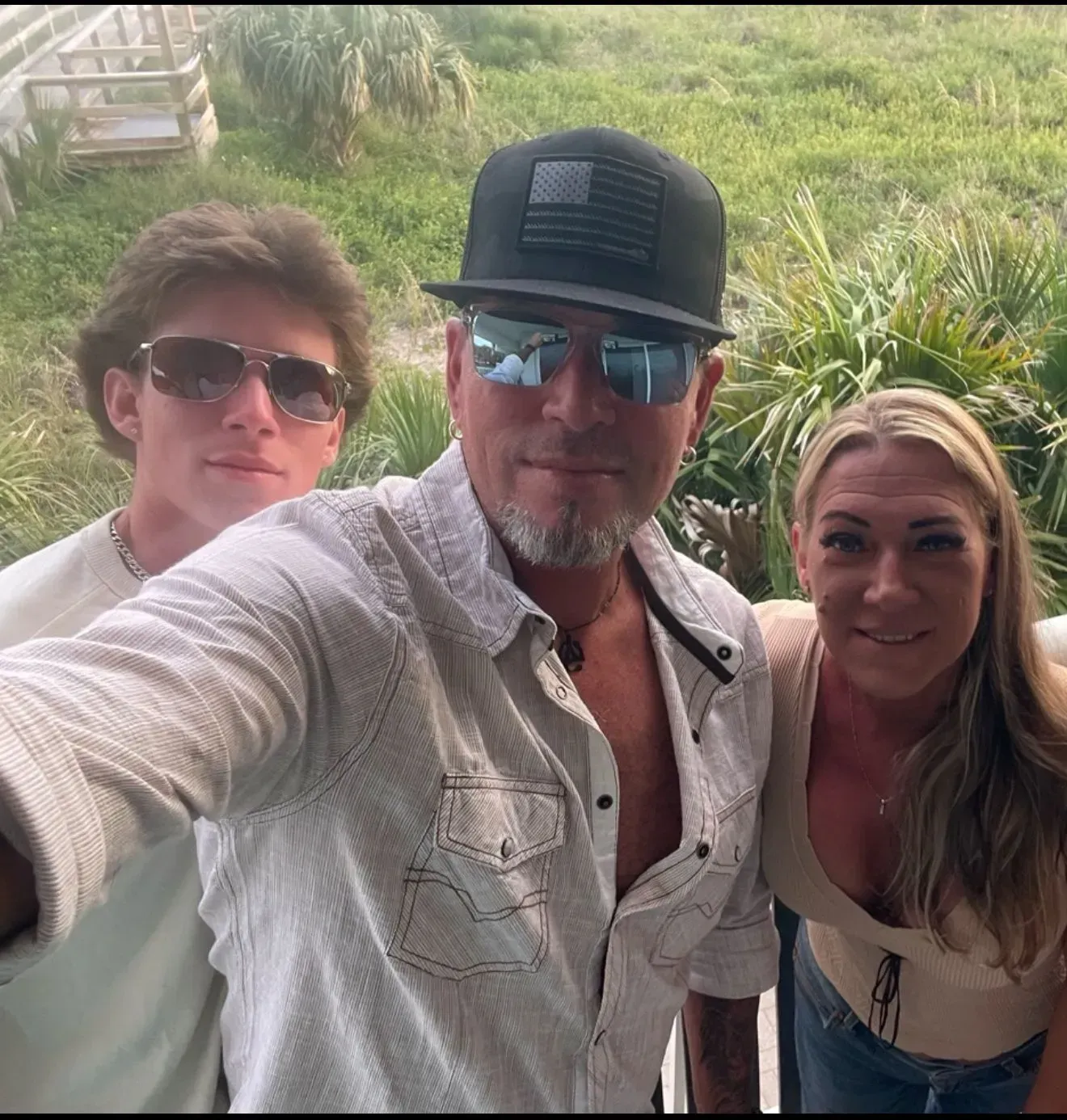 Three people smiling outside: a man in a cap, a teen with sunglasses, and a woman in a beige top.