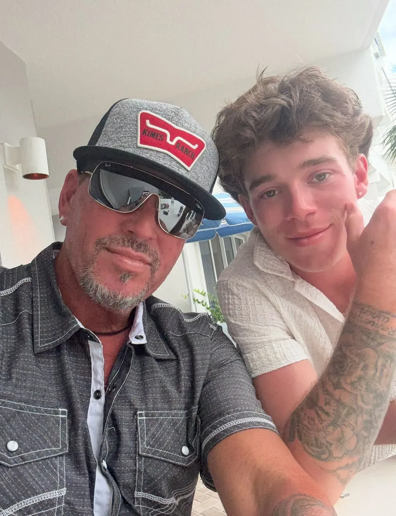 Man with sunglasses and cap next to a young man, both smiling. Outdoors.