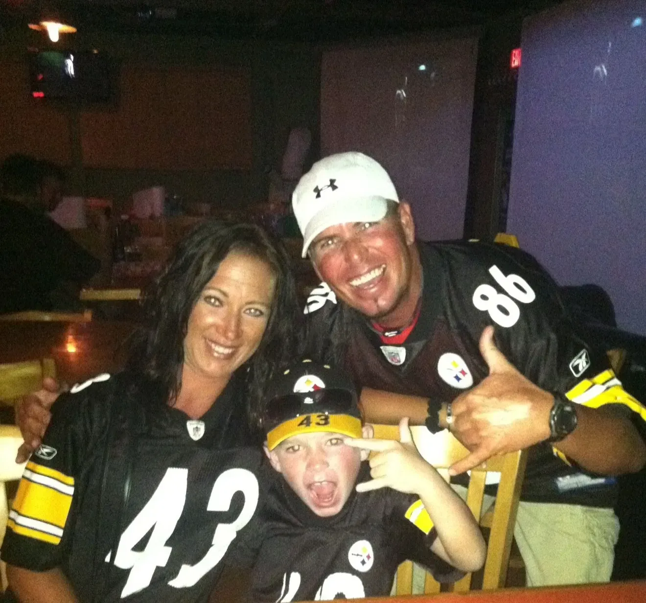 Family in Pittsburgh Steelers jerseys, smiling and making hand gestures in a dimly lit bar setting.