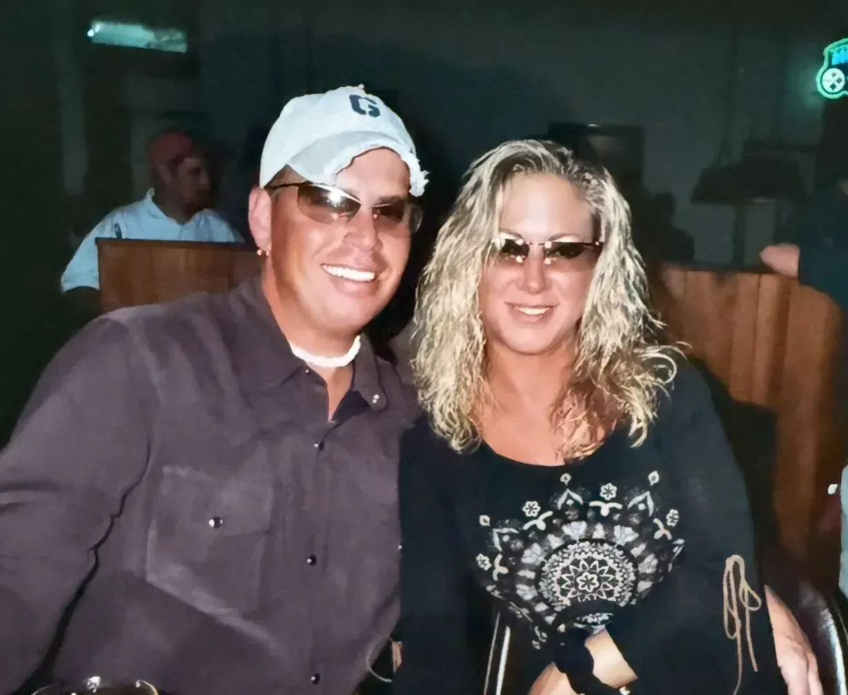 Smiling couple indoors; man in hat, sunglasses, brown shirt; woman in sunglasses, black top; wood paneling.