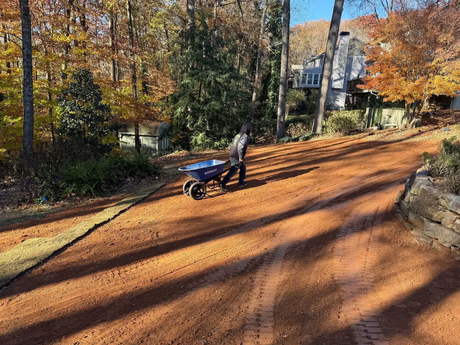 Person pushing a blue wheelbarrow on a reddish gravel driveway, surrounded by trees.