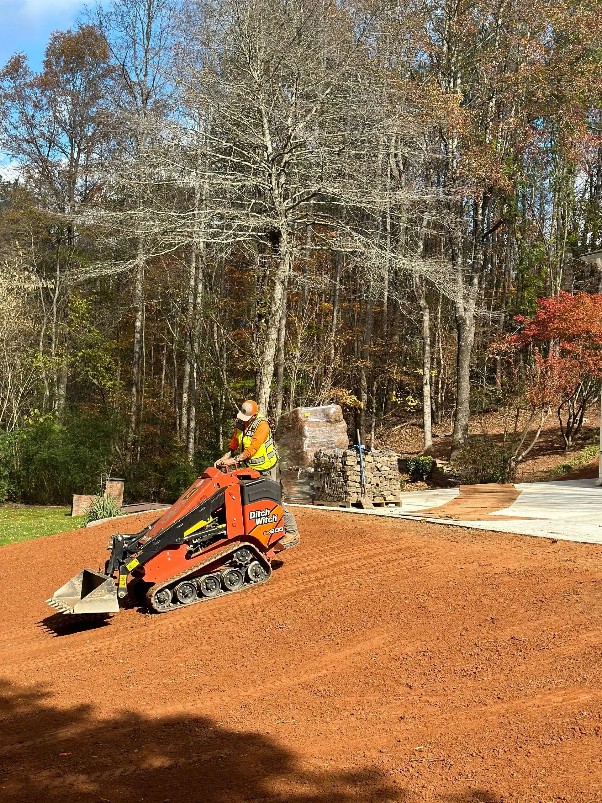 Mini excavator spreading red mulch on a lawn near a wooded area. Person operating the machine.