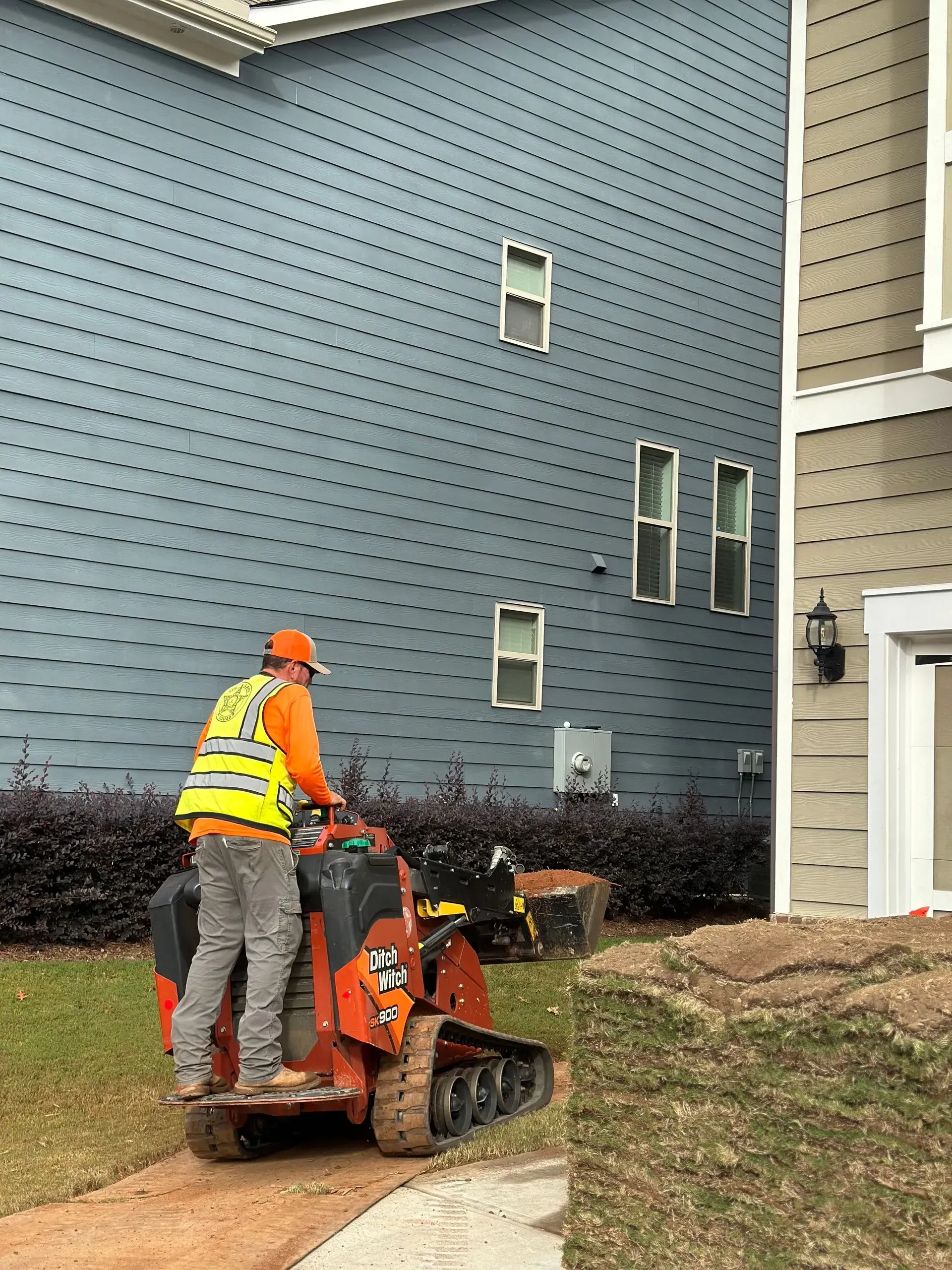 Worker operating a small orange skid steer near a blue building. Turf is stacked in the foreground.