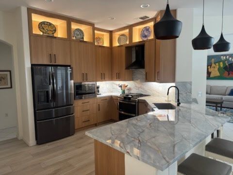 Modern kitchen with light wood cabinets, dark appliances, marble countertops, and pendant lights.