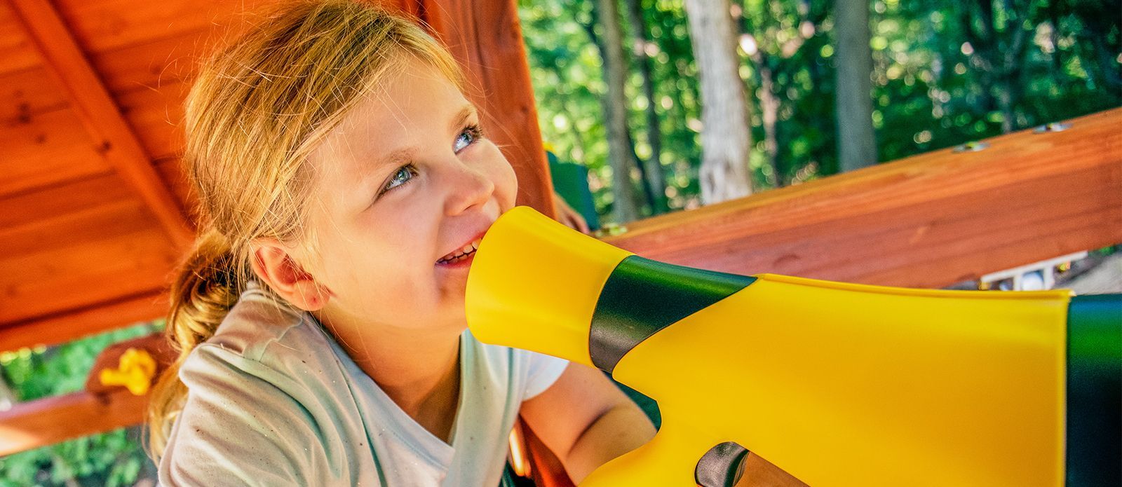 Child using a yellow and green horn-shaped toy under a wooden shelter, smiling outdoors