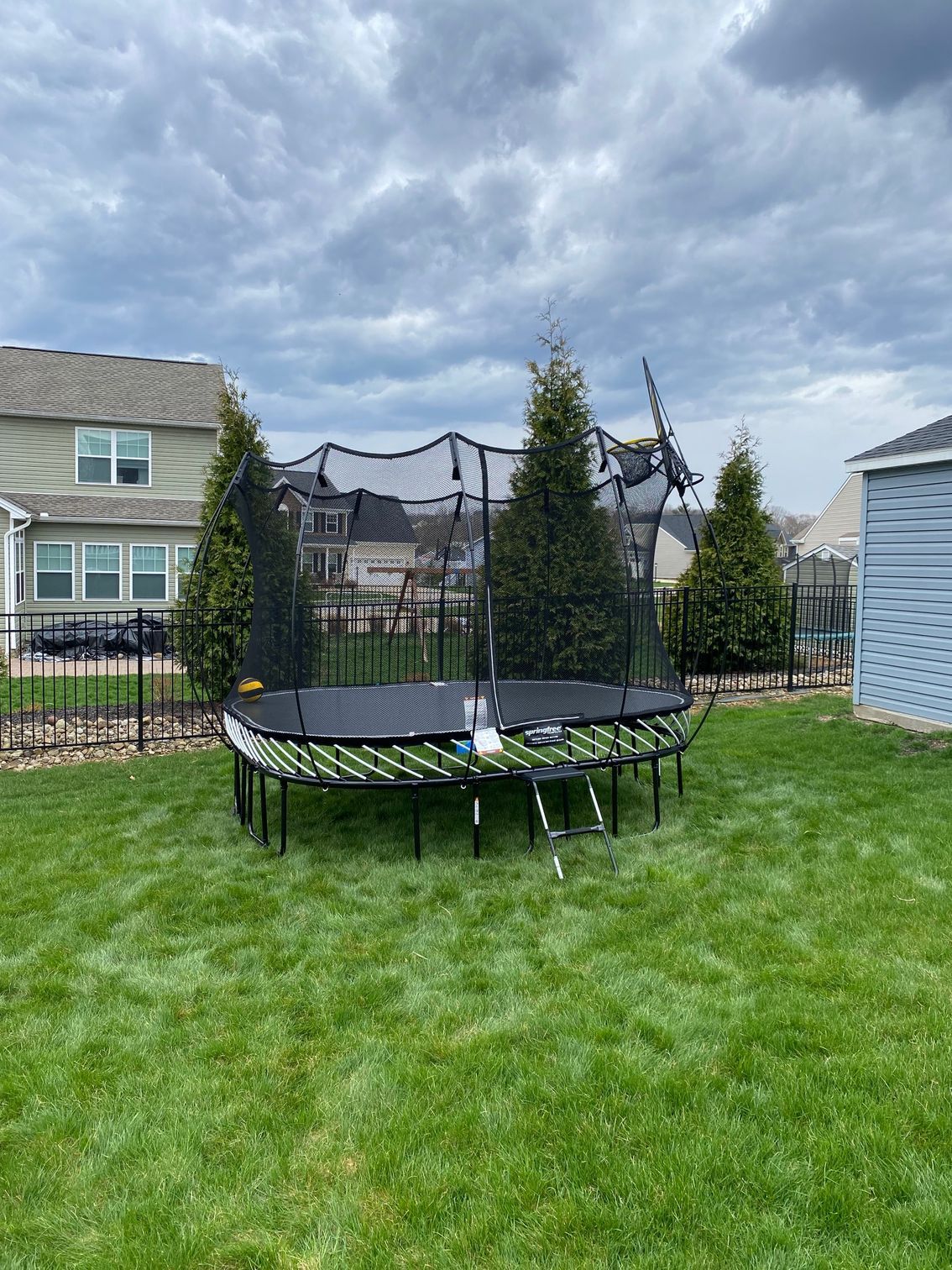 Round trampoline with safety net in a grassy backyard near houses under cloudy sky