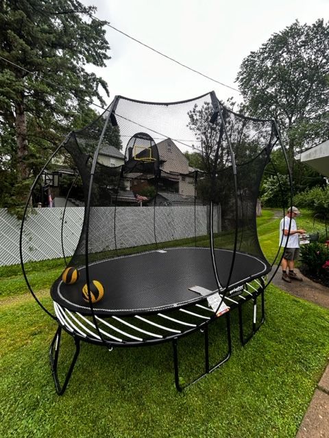 Backyard trampoline with safety net on a grassy lawn beside a white wall and trees