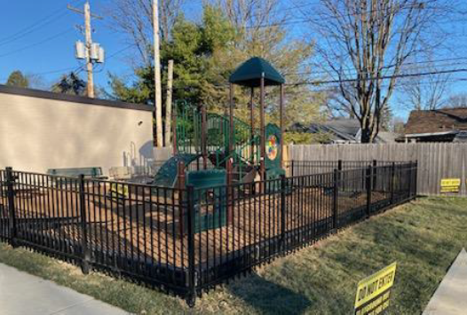 Small fenced playground with play structure and slides beside a building on a sunny day