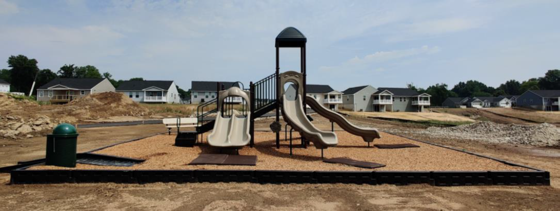 Playground with tan slides and climbing structures on a sandy lot under a partly cloudy sky